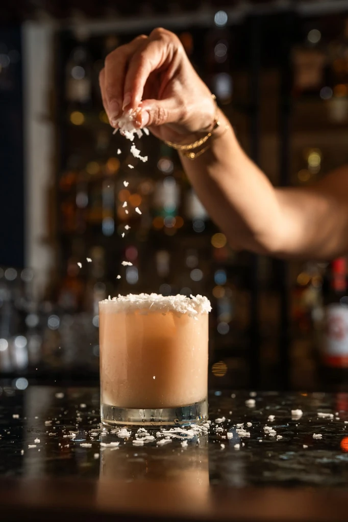 Hand sprinkling white garnish onto a peach cocktail at a bar.