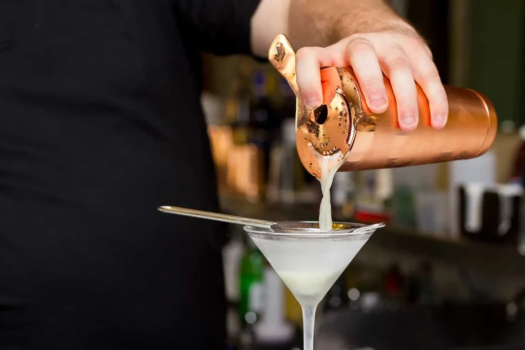 Bartender strains drink from copper shaker into frosted martini glass.