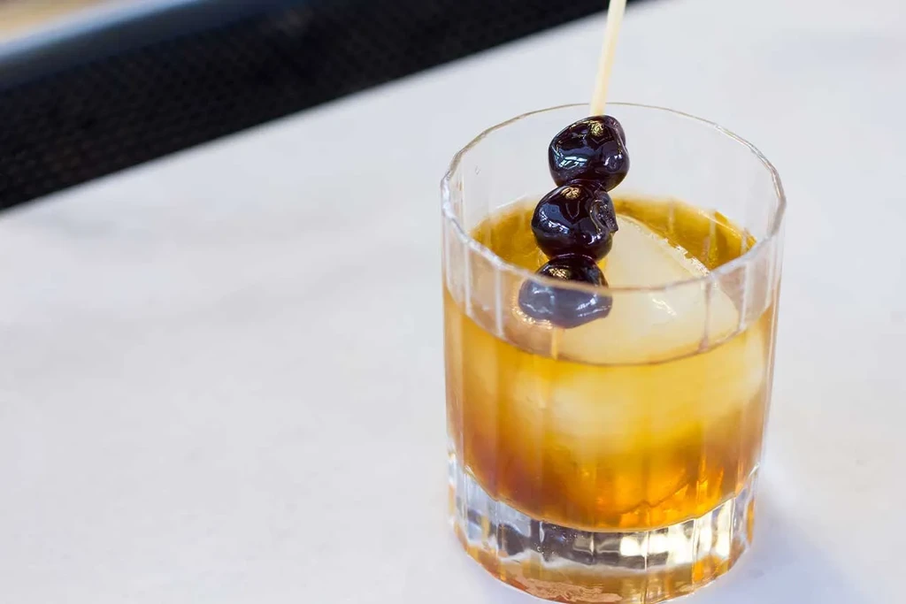 Amber cocktail in textured glass with spherical ice and cherry skewer.