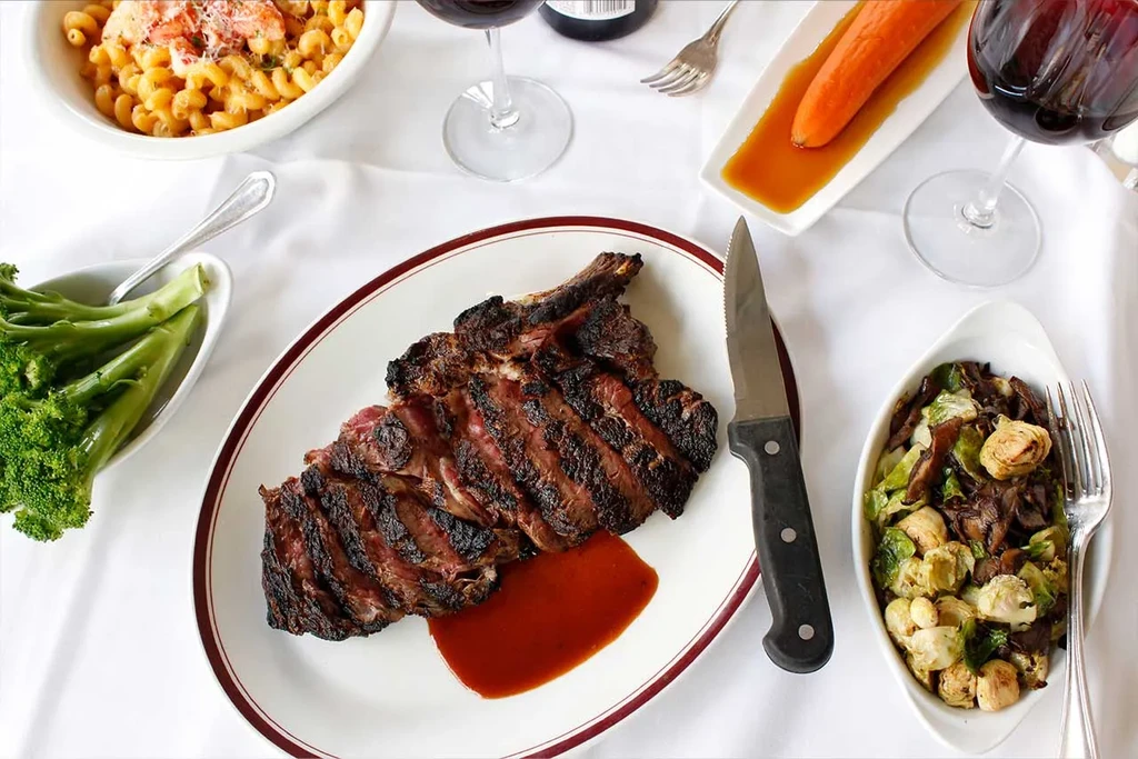 Pre-sliced bone-in steak, lobster mac and cheese, vegetables, and wine.