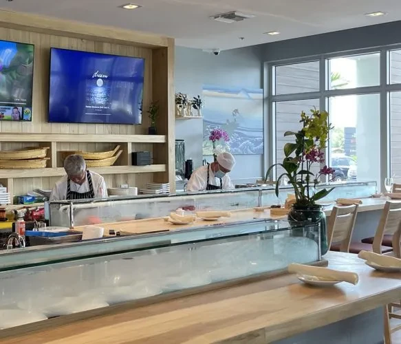 Two sushi chefs prepare food behind a modern wooden bar.