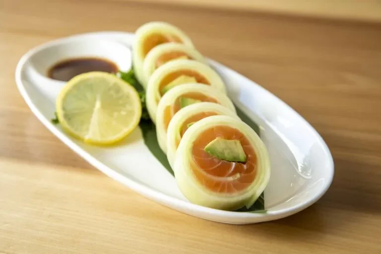 Cucumber-wrapped salmon and avocado rolls with dipping sauce and lemon slice.