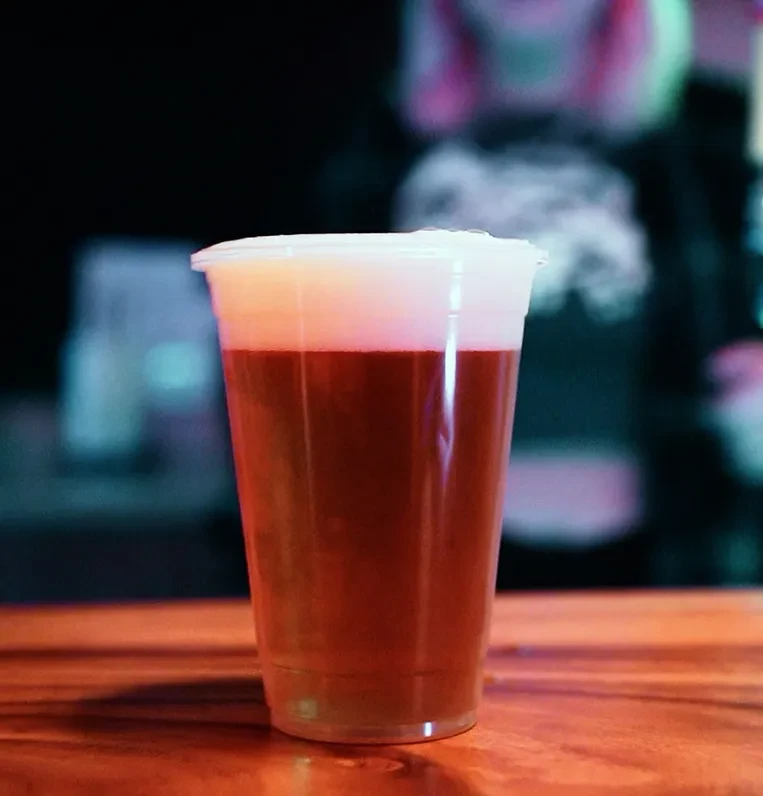 Plastic cup with layered reddish-brown drink and foam in a dark bar.