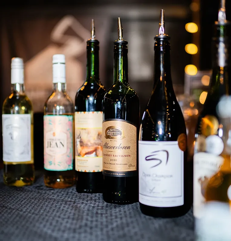 Multiple wine and spirit bottles with pour spouts on a dark bar.