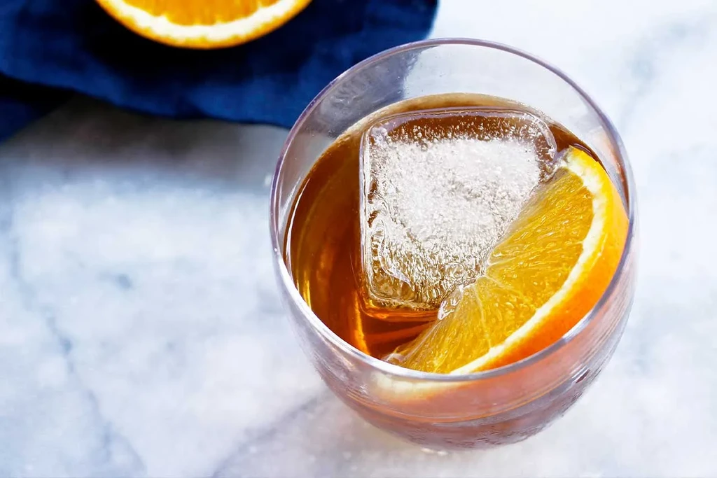 Overhead amber cocktail with orange slice and large ice cube on marble.