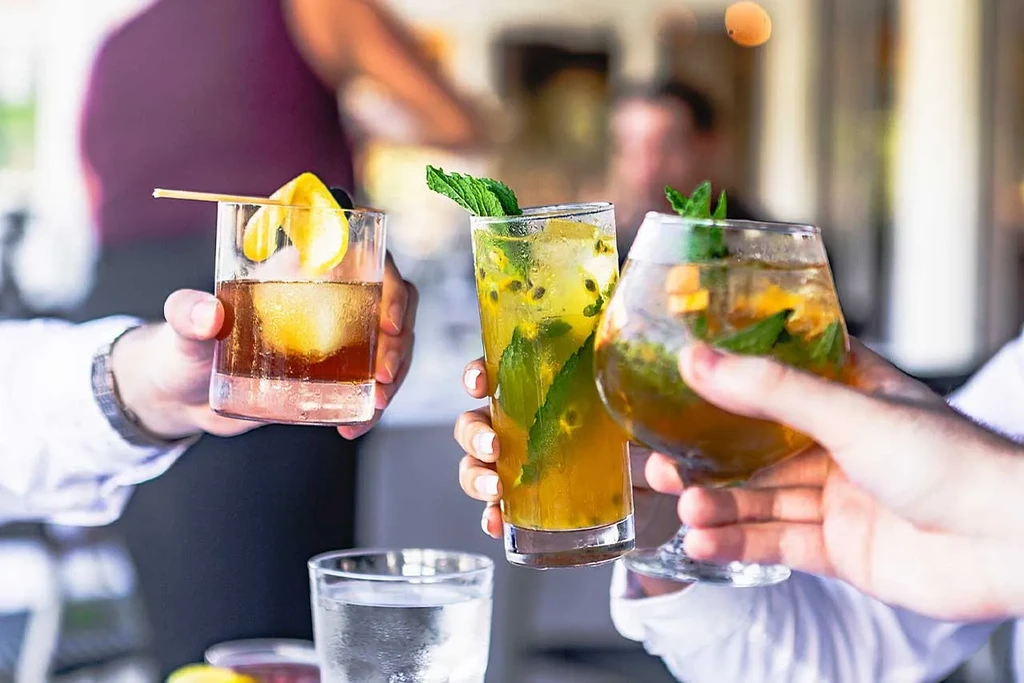 Three vibrant cocktails held by hands in an active bar setting.