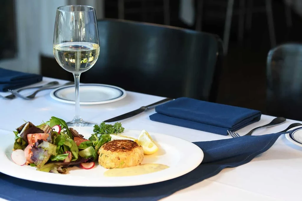 Plated crab cake, side salad, lemon, and white wine in restaurant.