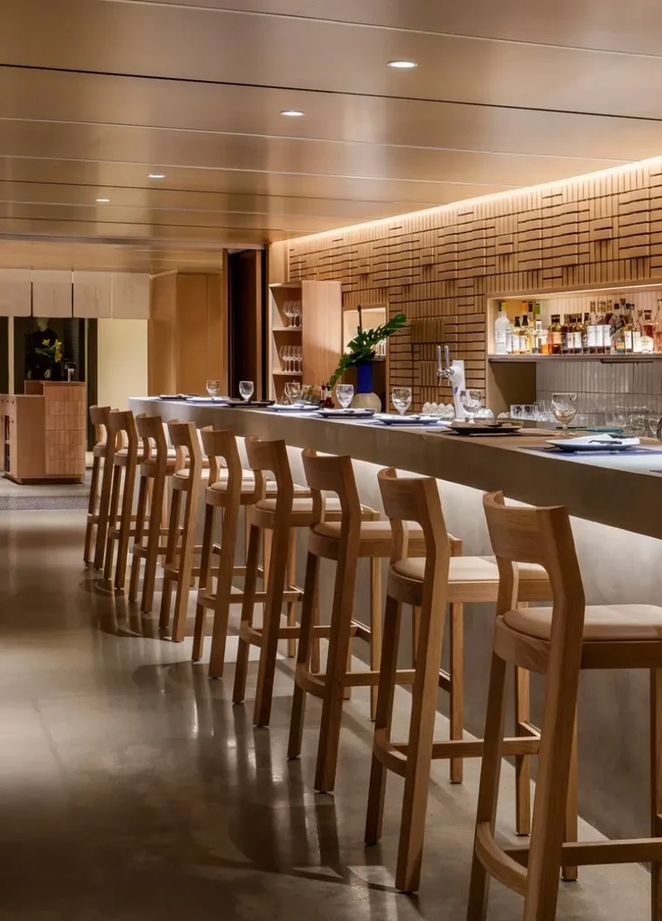 Warmly lit modern bar with long counter, wooden stools, and stocked shelves.