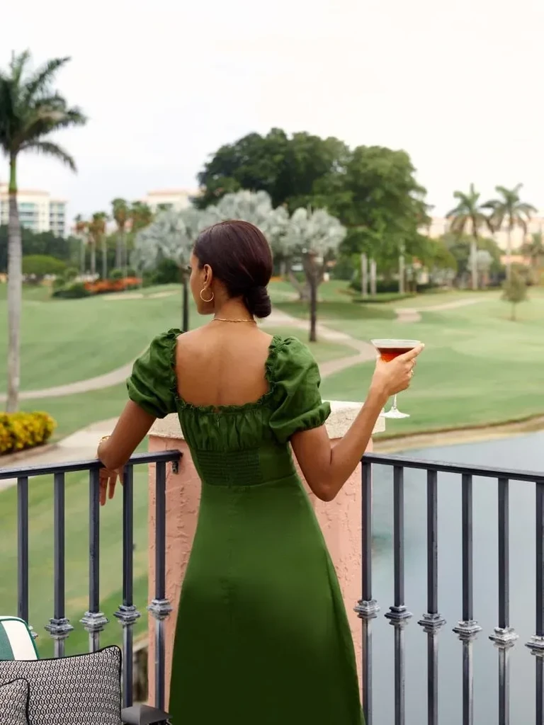 Woman in green dress holds cocktail, overlooking a resort golf course.