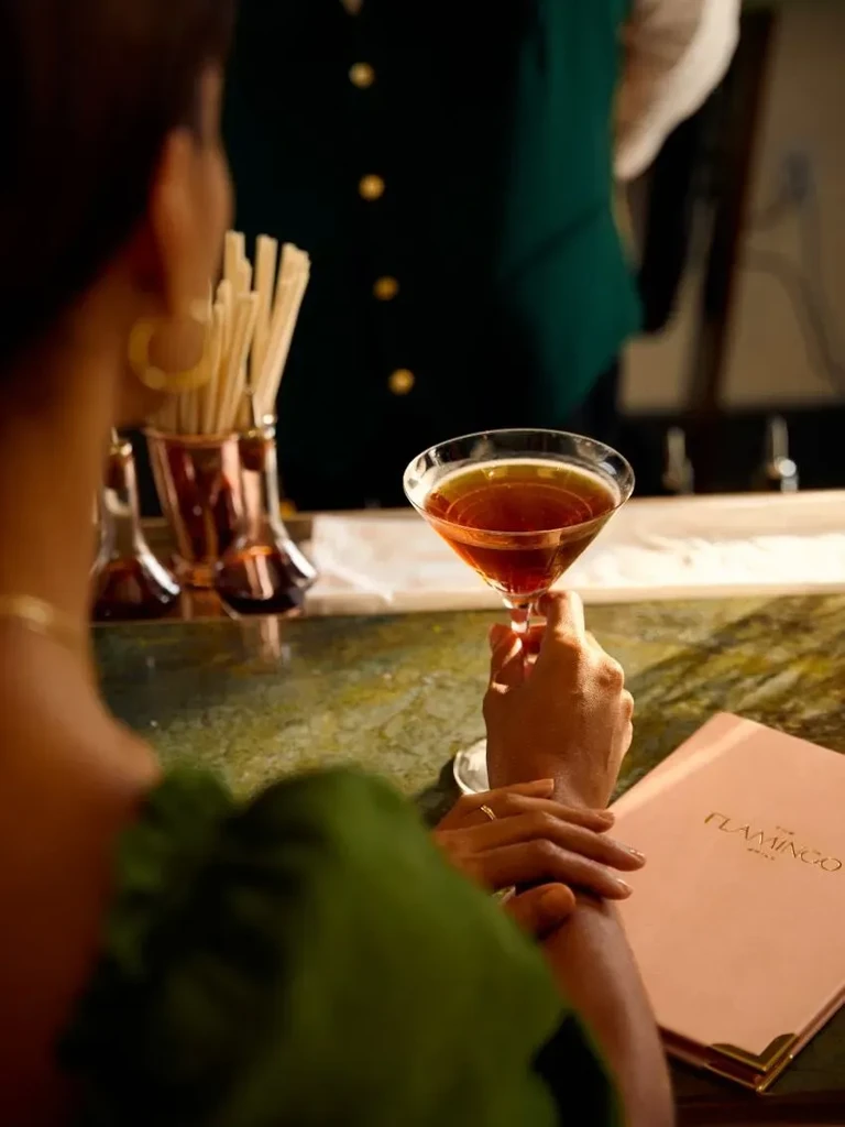Hands holding a reddish-brown martini next to 'THE FLAMINGO DRINKS' menu.