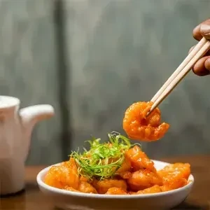 Hand lifting orange-glazed shrimp with chopsticks from a white bowl.