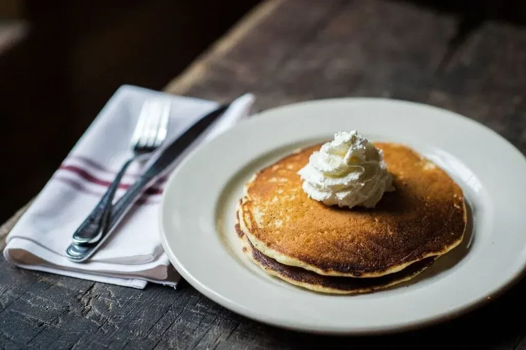 Stack of pancakes with whipped cream, cutlery, and striped napkin.