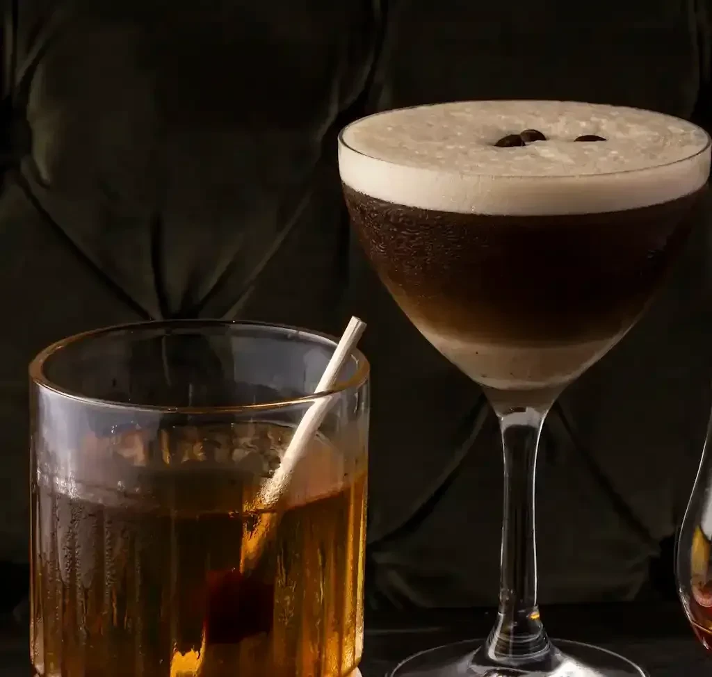 Layered Espresso Martini and amber spirit with cherry on dark fabric.