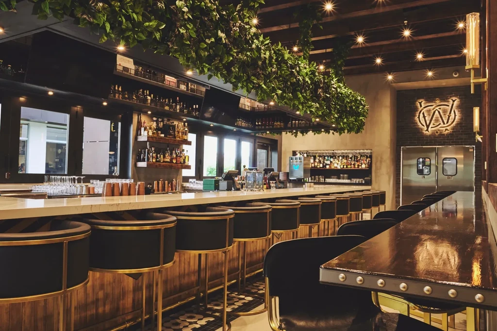 Modern, empty bar with stools, liquor bottles, lush greenery, warm lighting.