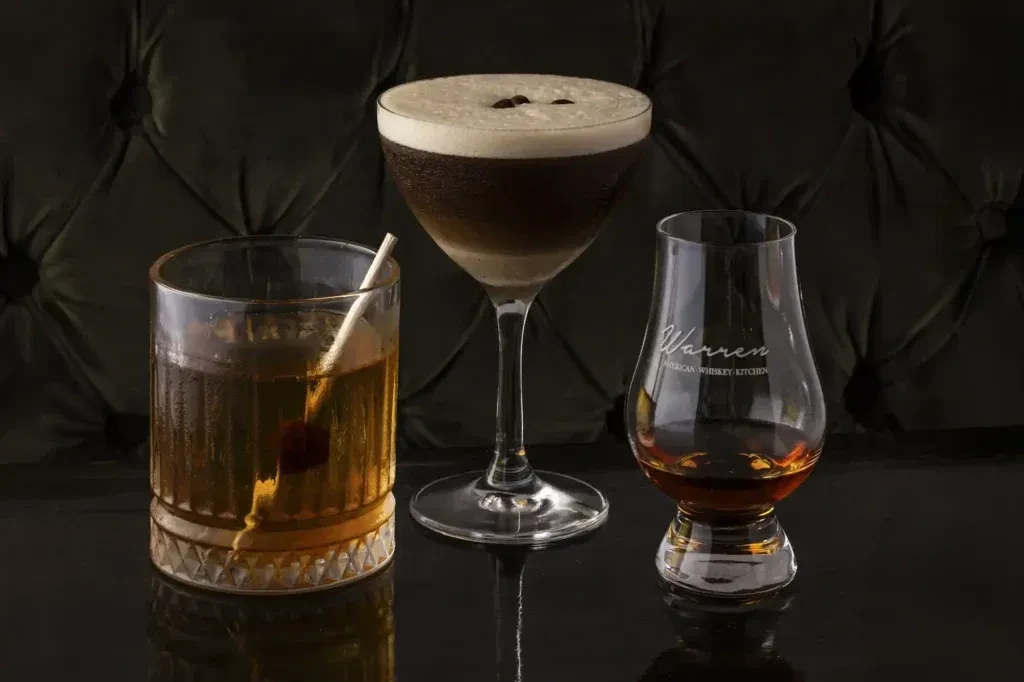 Three drinks: Warren whiskey snifter, layered cocktail, amber drink with ice.