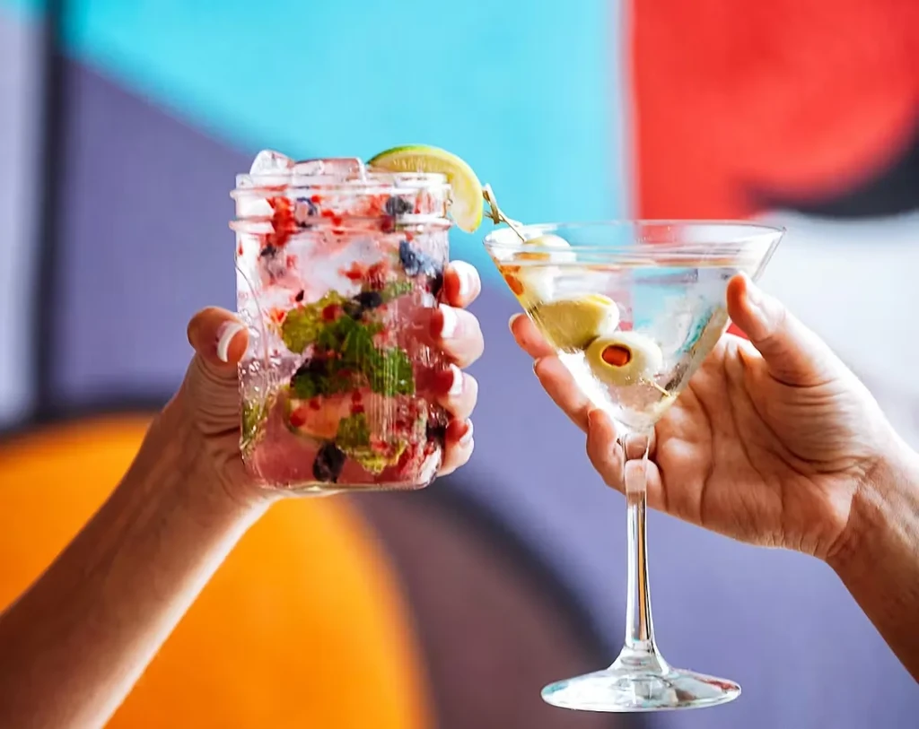 Hands hold a berry cocktail and martini against a colorful background.
