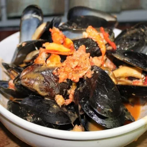 White bowl of cooked mussels in sauce with reddish topping.