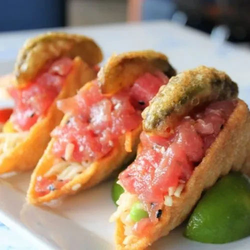 Three crispy tuna tacos topped with fried avocado and lime wedges.