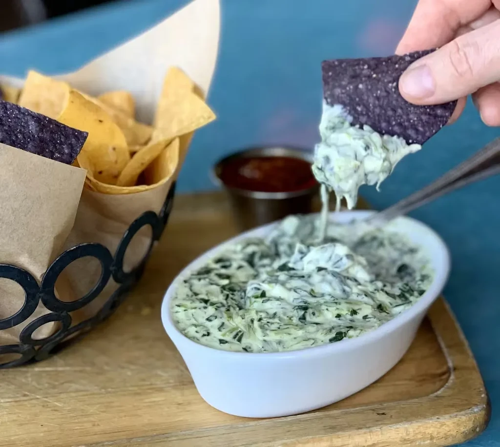 Hand dips blue corn chip into creamy green spinach artichoke dip.