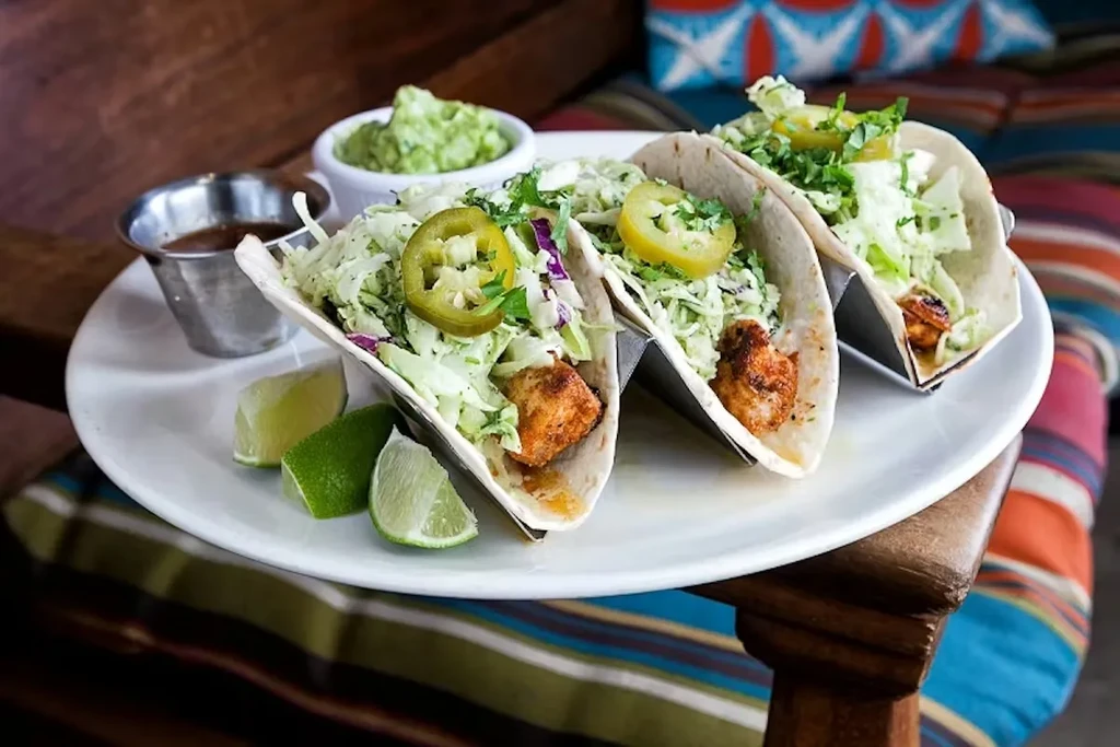 Three tacos in holders with fish, fresh toppings, lime, and salsa.