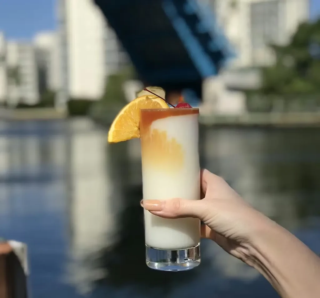 Layered cocktail with orange and cherry, held against a blurred city waterfront.