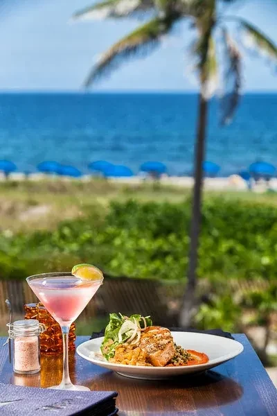 Plate of grilled fish, pink cocktail, tropical beach and ocean view.