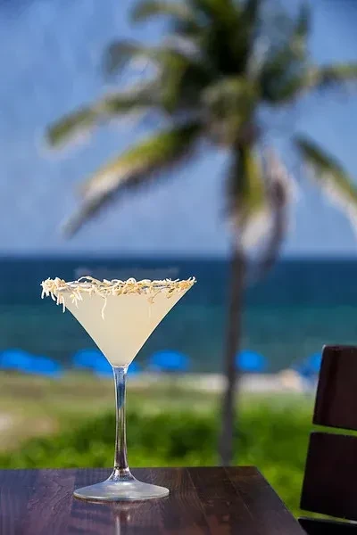 Coconut-rimmed martini on table, tropical beach and ocean background.