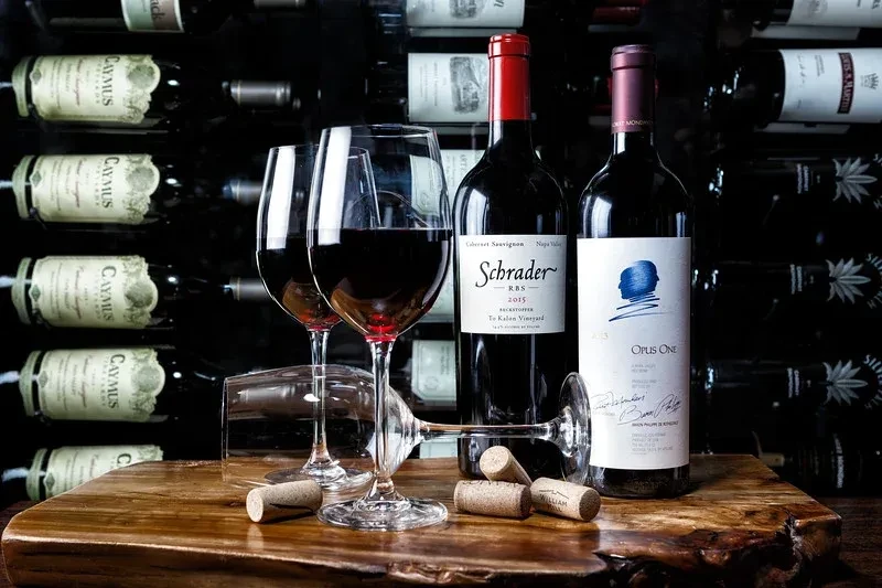 Schrader and Opus One red wines, glasses, and corks on wood.