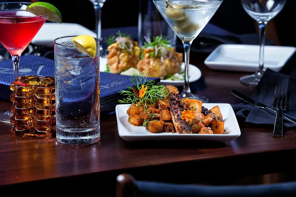 Grilled octopus dish, roasted potatoes, and three cocktails on a dark table.