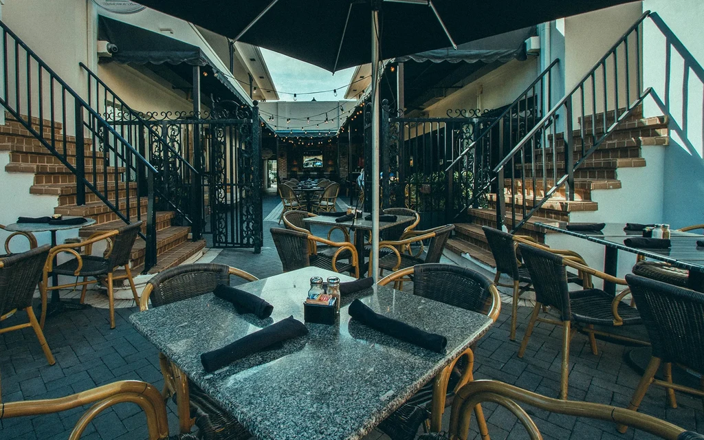 Courtyard restaurant patio with shaded tables, wicker chairs, and brick stairs.