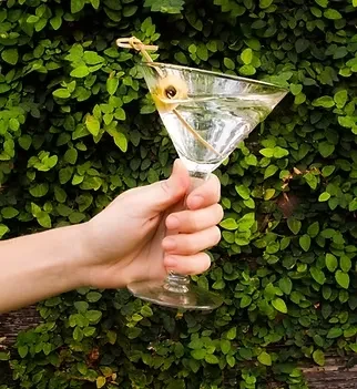 Hand holding martini glass with garnish against lush green foliage.