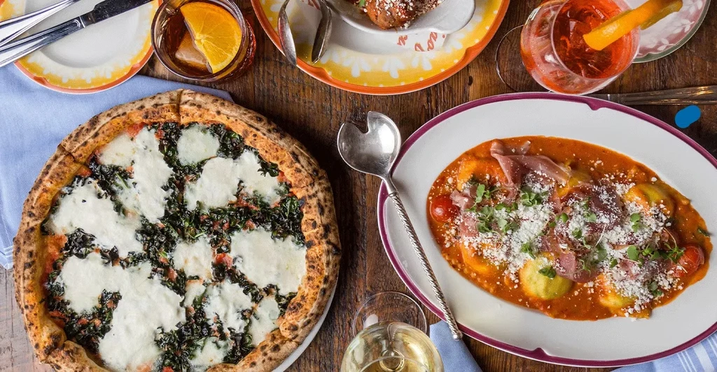 Overhead view of a wooden table with Italian pizza, pasta, and drinks.