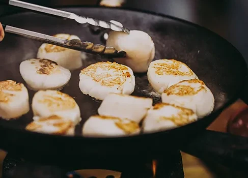 Large scallops searing in a dark pan with tongs.