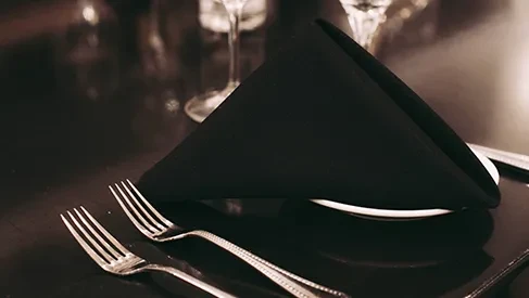 Low-light formal table setting with black napkin, white plate, and forks.