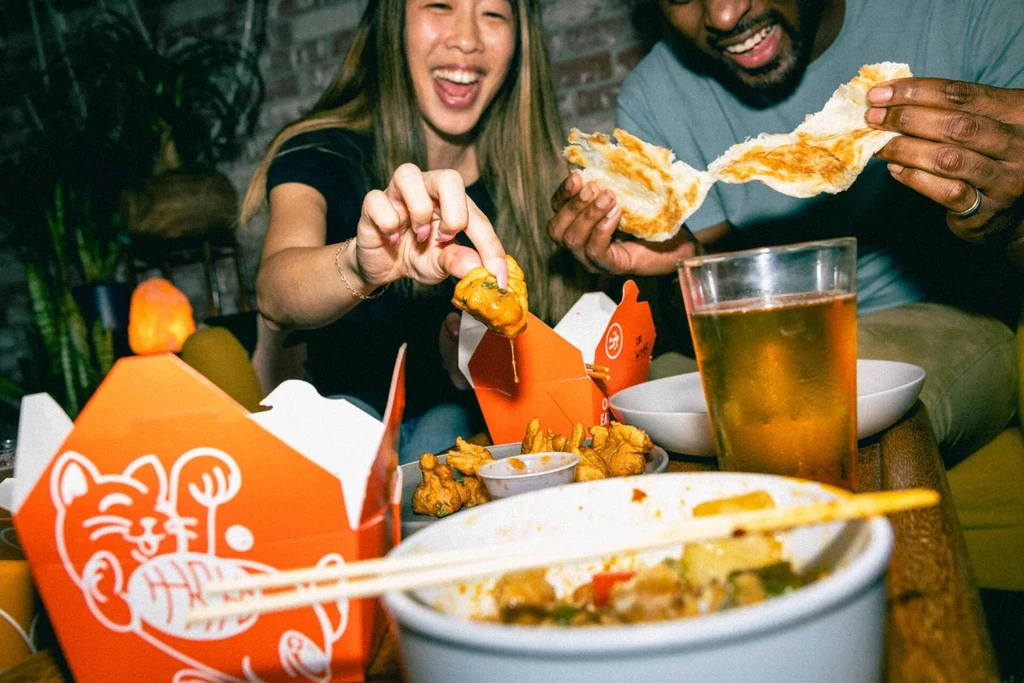 Man and woman smiling, sharing fried food, flatbread, and noodles.