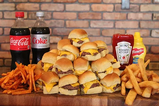 Mini cheeseburger sliders, fries, sodas, ketchup, and mustard on wood.