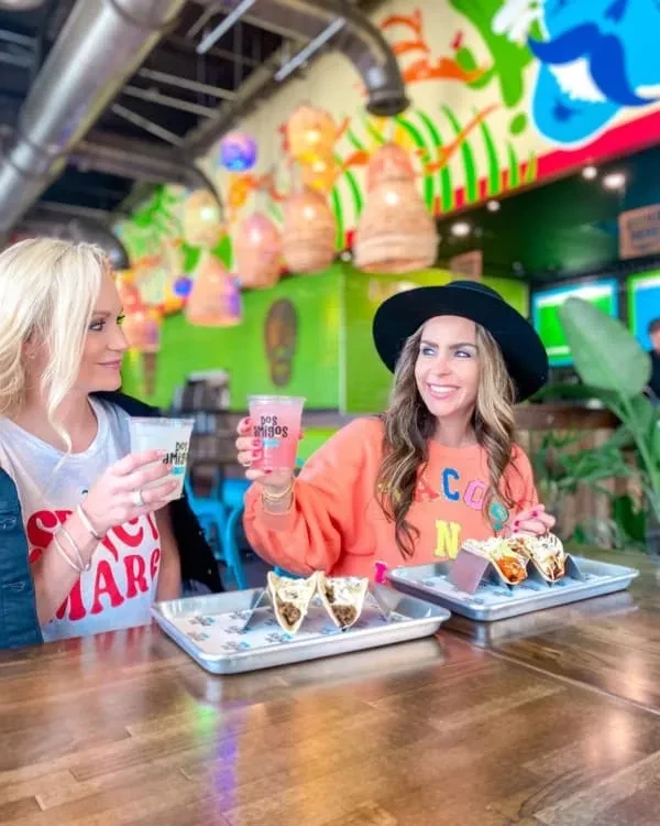 Two smiling women eat tacos and drink at a vibrant restaurant table.