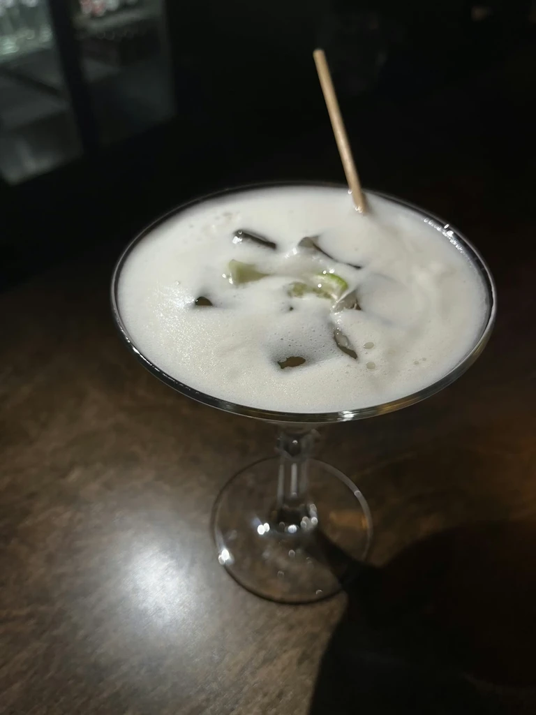 Cocktail in coupe glass with creamy white foam, lime, and ice.