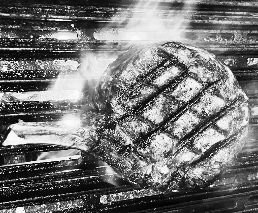 Black and white close-up of steak searing on grill with rising steam.