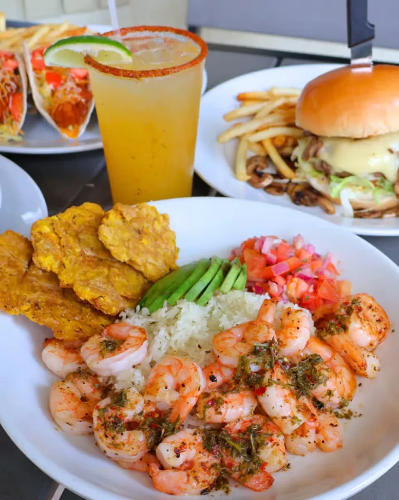 Restaurant table with shrimp, plantains, a cocktail, burger, and tacos.