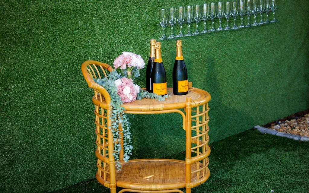 Rattan trolley with champagne, roses, and hanging flutes on turf.