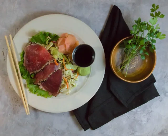 Seared tuna slices on a plate with ginger, wasabi, sauce, and chopsticks.