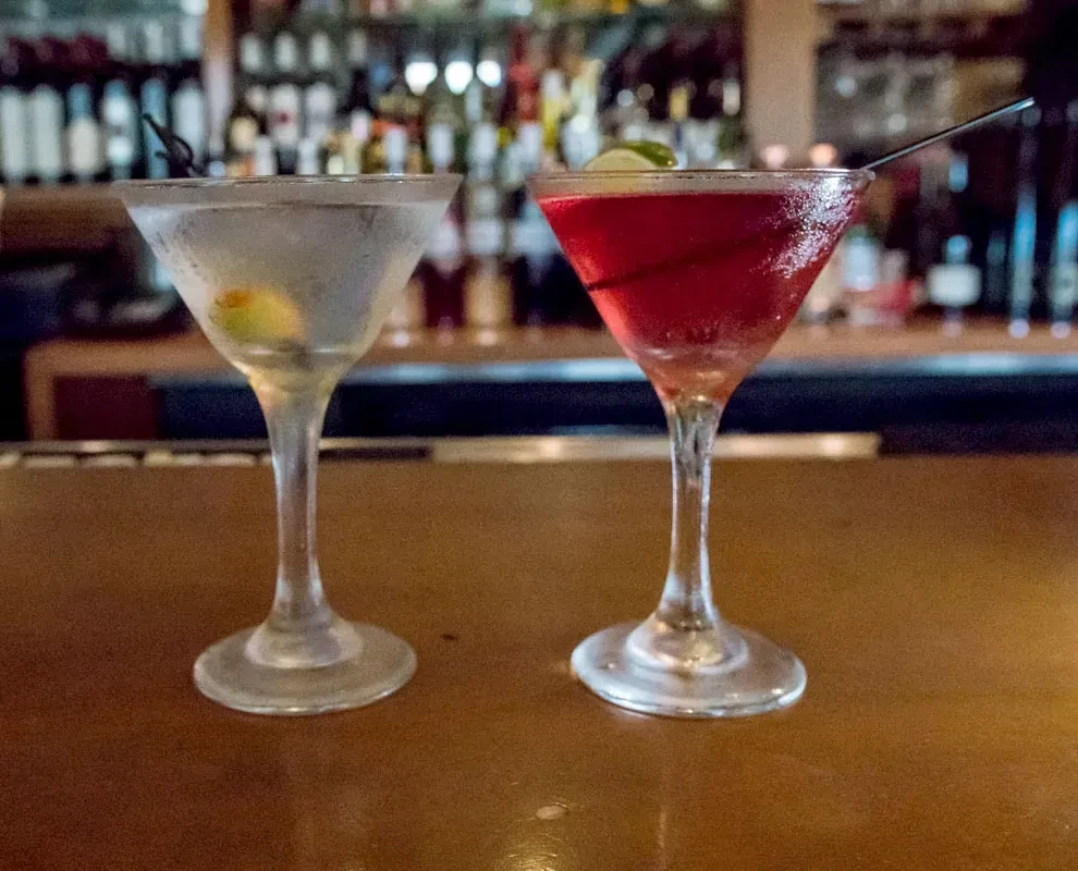 Two martini glasses with clear and red cocktails on a bar.