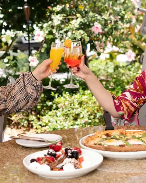 Hands toast with drinks, food, and hibiscus flowers on a patio.