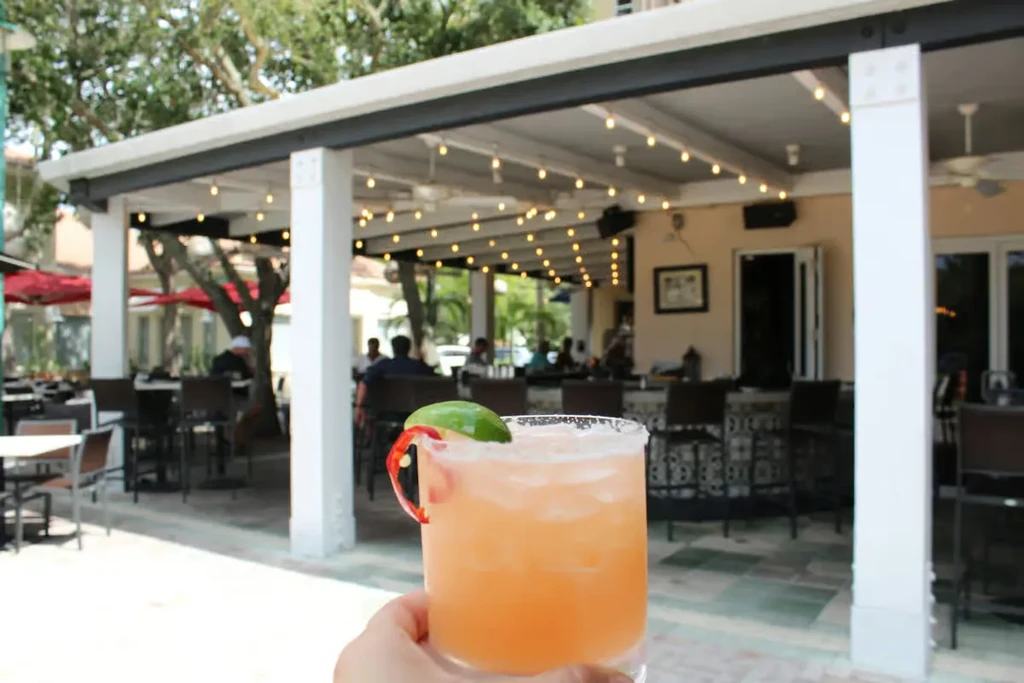 Hand holding an orange cocktail with lime and chili on a lively patio.