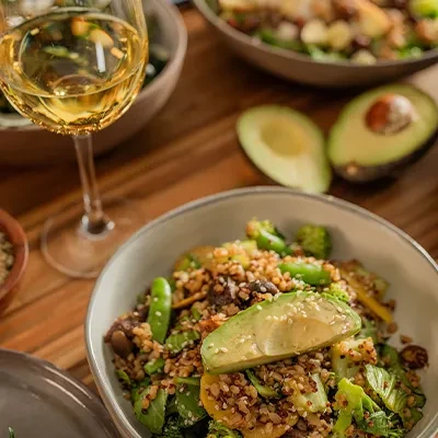 Grain salad bowl with avocado, vegetables, and wine on wooden table.