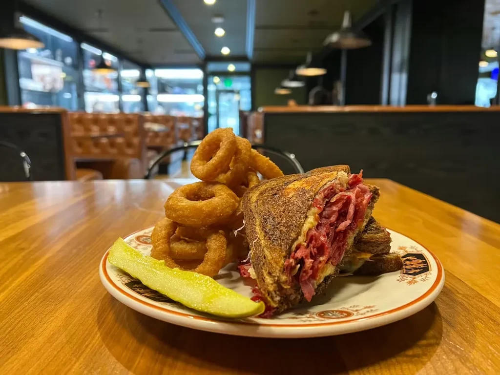 A large deli sandwich, onion rings, and pickle on a wooden table.