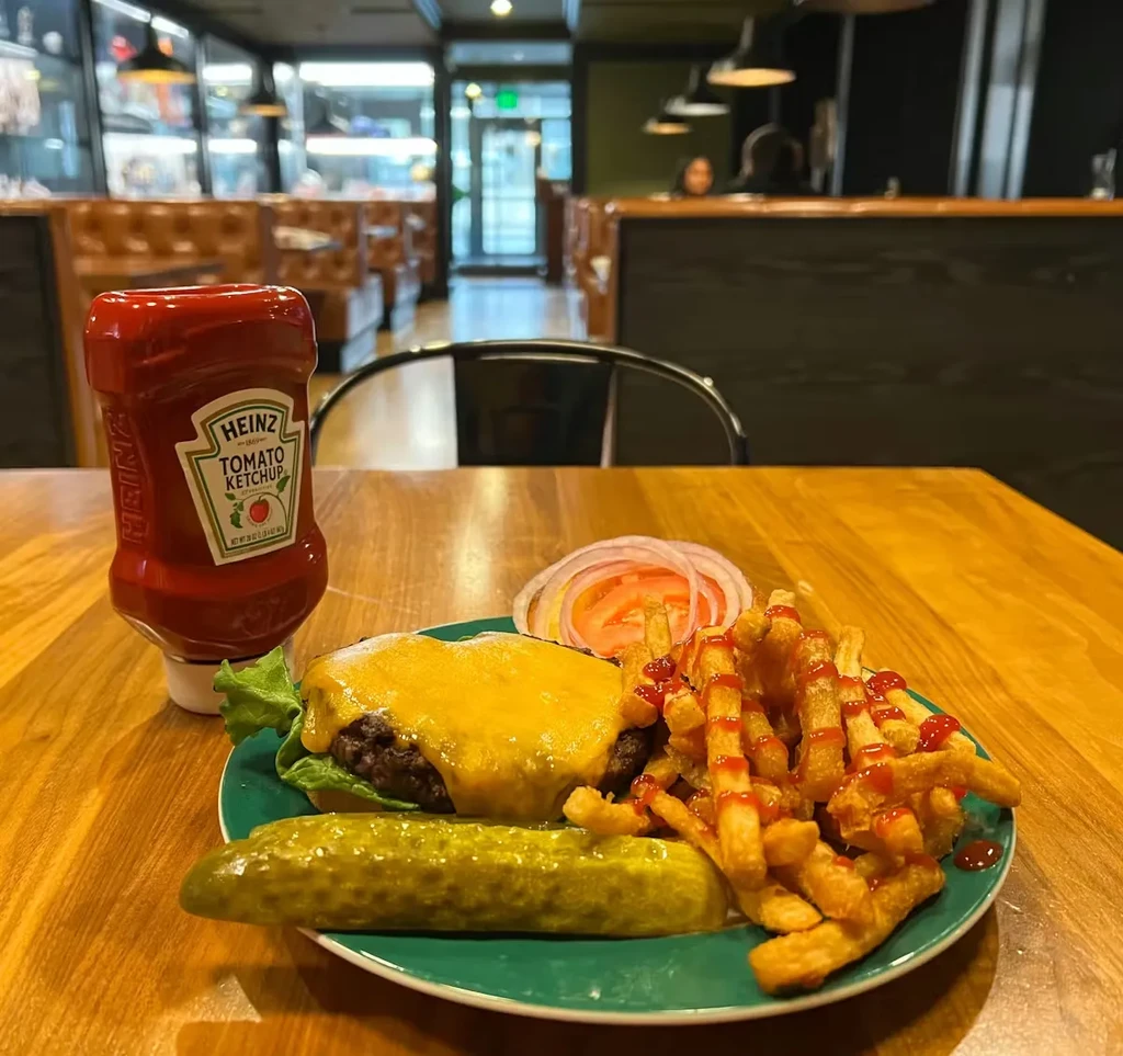 Cheeseburger, fries, pickle, and Heinz ketchup bottle on a diner table.