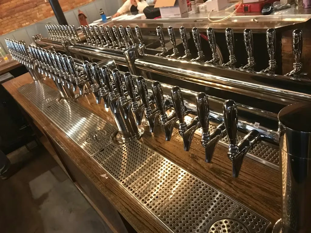 Numerous chrome beer taps with drip trays on a wooden bar.