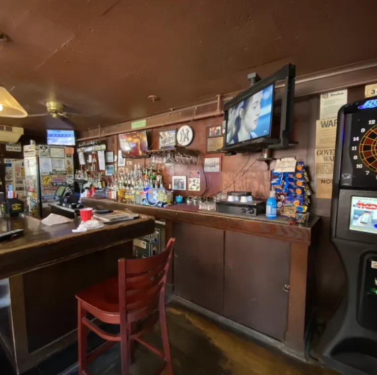 Dimly lit bar interior with stocked bar, TVs, and a dart machine.